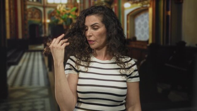 Woman covers mouth with hand and touches curly hair while seated in church building wearing striped shirt; serenity.