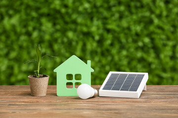 Portable solar panel with house figure, light bulb and plant on table outdoors. Green Energy concept