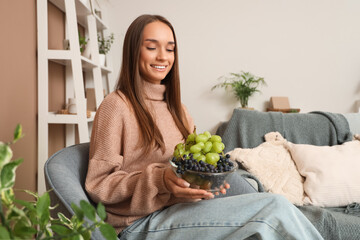 Young woman with grapes sitting in armchair at home