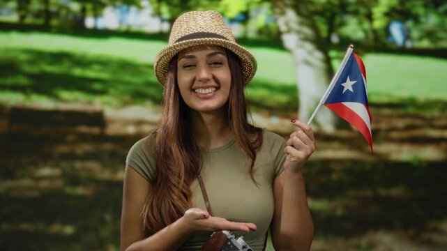 Woman smiling with puerto rican flag in park, young hispanic female holding camera outdoors in bright sun wearing straw hat and casual attire, representing travel and patriotism.