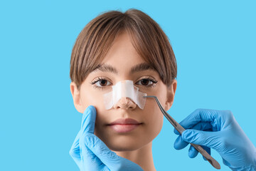 Surgeon hands with professional tool touching woman's bandaged nose after plastic surgery on blue...