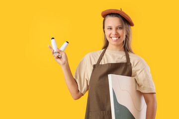 Young female artist with paints and painting on yellow background