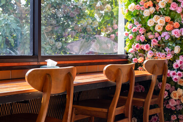A cozy cafe interior with wooden chairs at a counter, overlooking a window and a lush floral wall of pink and peach roses.