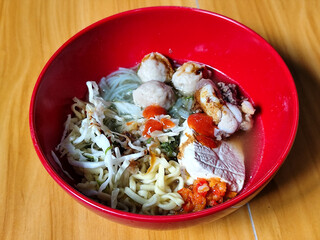 Bakso. The meatballs are served in a red bowl, containing meatballs, pieces of beef bone, yellow vermicelli noodles, vegetables, etc. Looks very delicious and delicious.