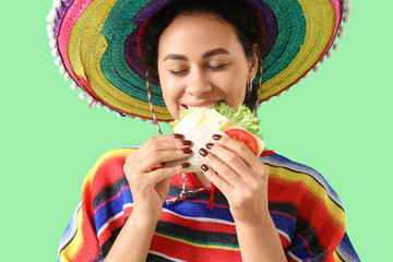 Young African-American woman in sombrero hat eating tasty taco on green background, closeup