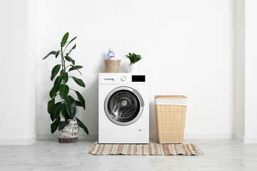 Modern washing machine with laundry basket and ficus houseplant near white wall in bathroom