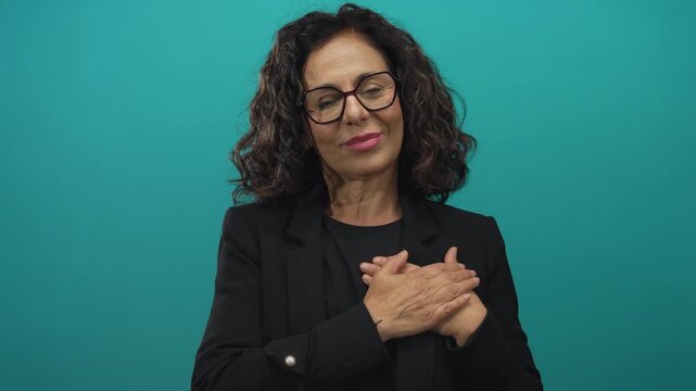 Woman wearing glasses in a black outfit stands with hands on chest against an isolated green background showing a peaceful and thoughtful expression.