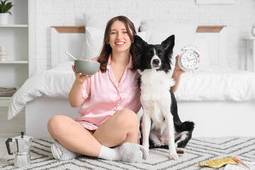 Beautiful young woman with sleep mask, cute Border Collie dog, breakfast and alarm clock sitting on...