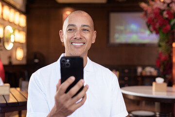 Confident Bald Hispanic man in white polo shirt sitting in Chinese style coffee shop using phone