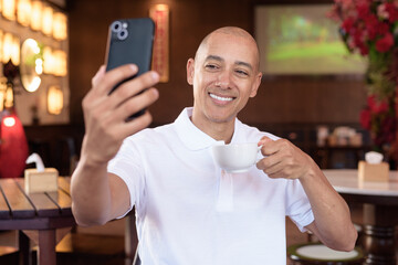 Confident Bald Hispanic man in white polo shirt sitting in Chinese style coffee shop using phone