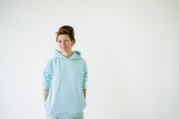 Studio portrait of a young woman with red hair wearing a light blue hoodie with hands in pockets,...