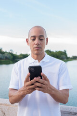 Confident bald man in white polo shirt using phone by lake in serene summer outdoor portrait