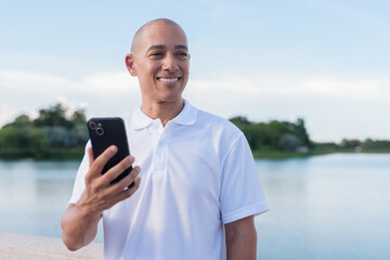 Confident bald man in white polo shirt using phone by lake in serene summer outdoor portrait