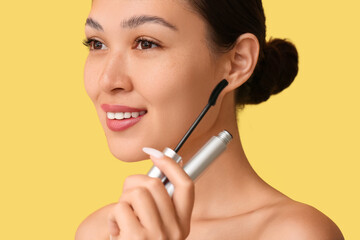 Happy young Asian woman with mascara on yellow background