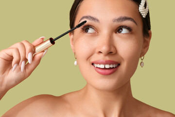 Happy young Asian woman applying mascara on green background, closeup