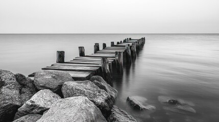 black and white image of an old wooden pier extending into calm water with large rocks in the foreground under a clear sky