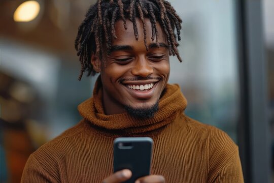 young man with dreadlocks wearing brown turtleneck sweater smiling while looking at smartphone outdoors
