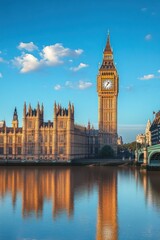 Obraz premium Historic gothic-style clock tower and surrounding parliamentary building illuminated by warm sunlight reflecting on calm river under clear blue sky with scattered clouds