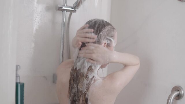 Blond woman lathering hair during slow motion shower, enjoying streaming water inside sleek minimalist bathroom with bright lighting and clean surfaces