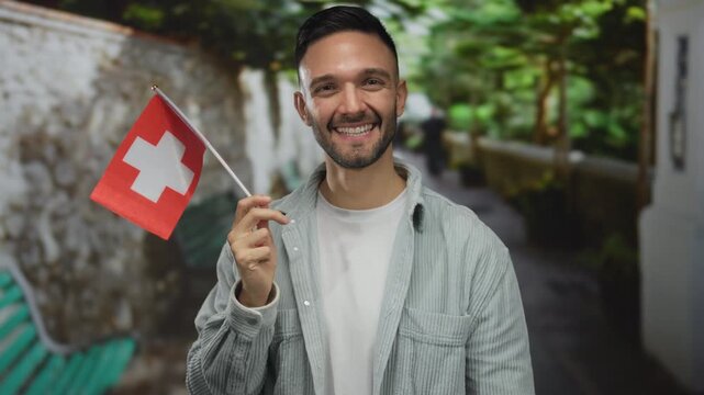 Young hispanic man smiling and holding a swiss flag outdoors in a green park setting, showcasing his casual attire and cheerful expression.