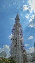 A towering mosque minaret under renovation, surrounded by scaffolding, rises into a bright blue sky as sun rays filter through scattered clouds, creating a majestic view.