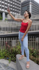 A smiling young Asian woman in a red top and blue jeans smiles while taking a selfie in an urban...