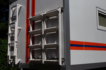 Close up of side of emergency rescue vehicle showing mounted ladder and storage racks. The bright orange and blue stripes, along with functional design, emphasize its role in emergency operations