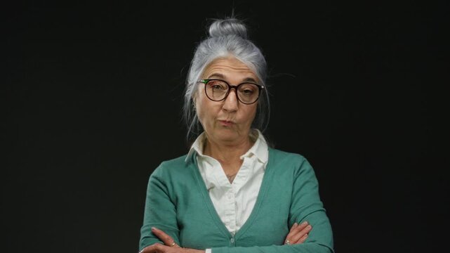 Senior grey-haired woman with glasses posing against a black background wall, expressing a contemplative mood with her arms crossed.