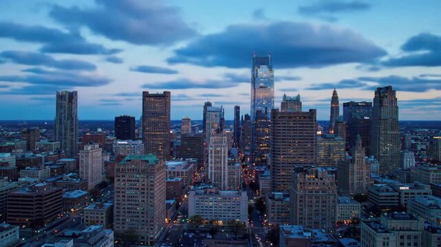 An aerial view showcases a vibrant cityscape with numerous skyscrapers reaching towards a cloudy, colorful sky during the day.