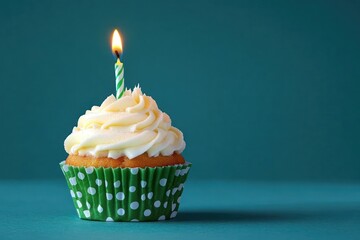 Single cupcake with white frosting and a lit candle in a green polka dot wrapper on a teal background