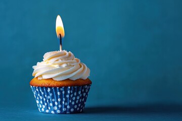 Single cupcake with white frosting and lit candle in blue polka dot wrapper against plain blue background evoking celebration and simplicity