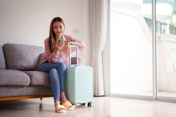 Asian traveler woman happily uses smartphone plans exciting journey home comfortable modern living room for upcoming vacation trip
