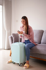 Asian adult woman happy plans trip using smartphone beside suitcase at home prepare for vacation...