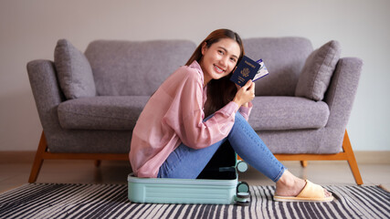 Asian young adult woman excitedly prepares travel vacation holding passport boarding pass smiling inside suitcase home living room adventure trip concept