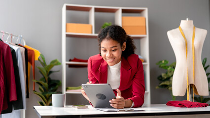 African American Fashion Designer Using Tablet for Online Business and Design at Her Stylish Workspace with Mannequin Displaying Her Newest Creations