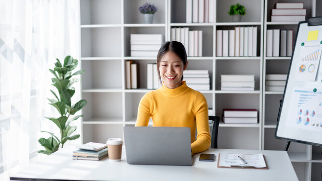 Young Asian Woman Working From Home on Laptop Computer Smiling and Confident. Home Office Setup with Bookshelf and Financial Report Board. Remote Work and Online Communication