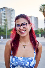 Portrait of a cheerful young woman with red hair and glasses smiling in a city environment, looking...