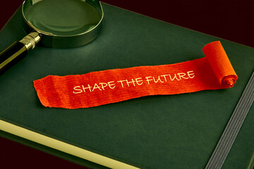 A notepad lies on a dark surface, featuring the motivational phrase Shape the Future written on an...