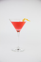 A red cocktail in a classic martini glass with a twist of orange peel set against a white background