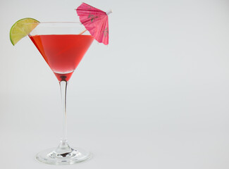 A red cocktail in a classic martini glass with a wedge of lime and an umbrella set against a white background.