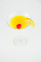 An orange cocktail in a classic martinin glass with a wedge of lemon and a red cocktail cherry set against a white background viewed from above.