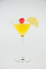 An orange cocktail in a classic martinin glass with a wedge of lemon  and a red cocktail cherry set against a white background.