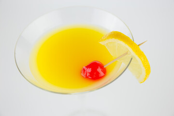 An orange cocktail in a classic martinin glass with a wedge of lemon and a red cocktail cherry set against a white background viewed from above. This a macro image with selected focus.