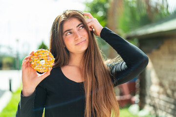 Young pretty caucasian woman holding a donut at outdoors having doubts and with confuse face...