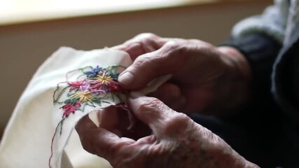 A close-up view shows hands delicately working with needle and thread, meticulously crafting a floral design onto a piece of fabric.