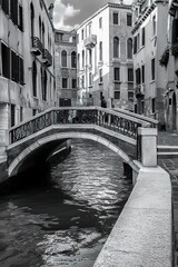 Fototapeta premium black and white photo of a narrow canal with a stone arch bridge and old multi-story buildings on both sides under a partly cloudy sky