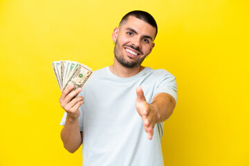 Young caucasian man taking a lot of money isolated on yellow background shaking hands for closing a...