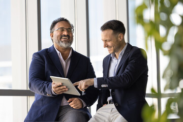 Two cheerful diverse mature and young professional men discussing creative ideas for online product development, using digital tablet, pointing at gadget, laughing, enjoying teamwork