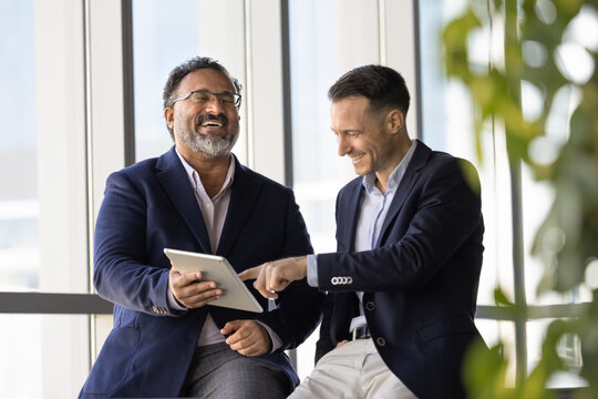 Two cheerful diverse mature and young professional men discussing creative ideas for online product development, using digital tablet, pointing at gadget, laughing, enjoying teamwork