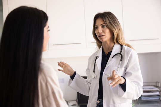 Serious doctor woman in lab coat talking to female patient in hospital examination room, giving medical recommendation for treatment, therapy, telling diagnosis, checkup result - Powered by Adobe